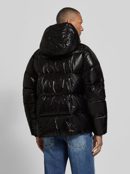 Męski model nosi kurtkę Regular Fit Jacke mit Kapuze Modell '3D Down' od Dsquared2, czarny połysk, krój regular fit, kaptur z tunelem, zamek błyskawiczny dwustronny, boczne kieszenie zapinane na zamek, prążkowane zakończenia rękawów, długość tyłu w rozmiarze 50: 75 cm, szerokość ramion w rozmiarze 50: 51 cm, szerokość klatki piersiowej w rozmiarze 50: 64 cm, długość rękawów w rozmiarze 50: 68 cm, widok całej sylwetki z tyłu.
