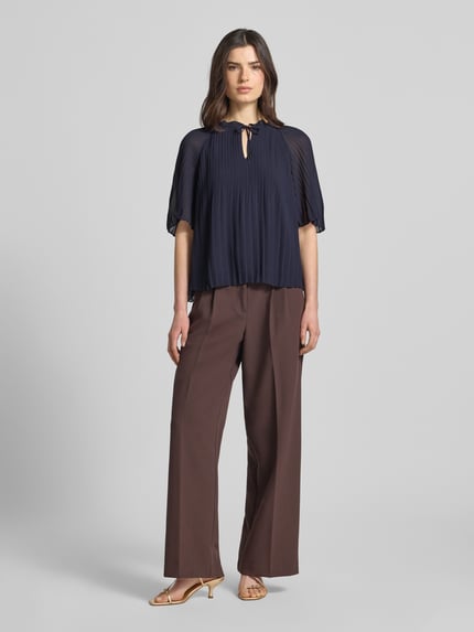 Frauenmodel trägt Regular Fit Blusenshirt in Plissee-Optik von s.Oliver BLACK LABEL in Dunkelblau, Optik: Unifarben, Passform: Regular-Fit, Rüschenkragen, Plisseefalten, Rundhals, Schlüsselloch, Schnürung, Ärmelform: Kurzarm, glatte Struktur, weite braune Hose, Ganzkörperansicht von vorne.