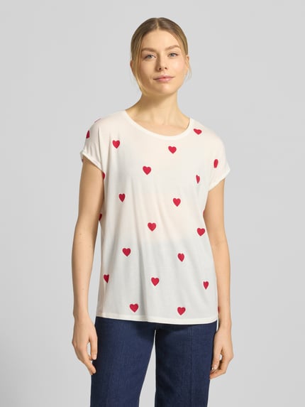 Kobiece model nosi Relaxed Fit T-Shirt aus Lyocell-Mix Modell 'AVA' od Vero Moda, efekt: wzór na całej powierzchni, nadruk na całej powierzchni, okrągły dekolt, krótki rękaw, zakończenie rękawów: brzeg, materiał: mieszanka lyocellu, jasny kolor z czerwonym wzorem serc, gładka faktura, widok całej sylwetki z przodu.