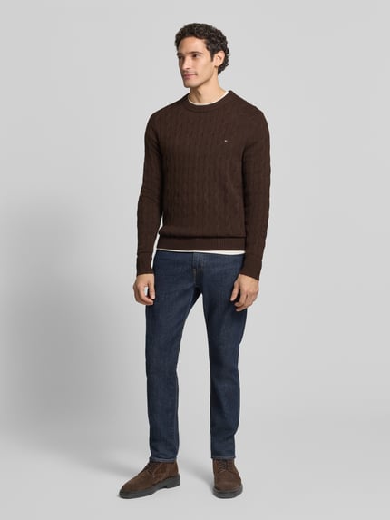 Männermodel trägt Regular Fit Pullover aus reiner Schafswolle von Tommy Hilfiger in Braun mit Zopfmuster, Longline, Logo-Detail, Ganzkörperansicht von vorne.