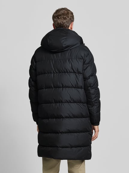 Männermodel trägt Steppmantel mit Kapuze Modell 'H-Carpa' von BOSS in Schwarz, Longline, mit Label-Detail und Taschen, Ganzkörperansicht Rückansicht.
