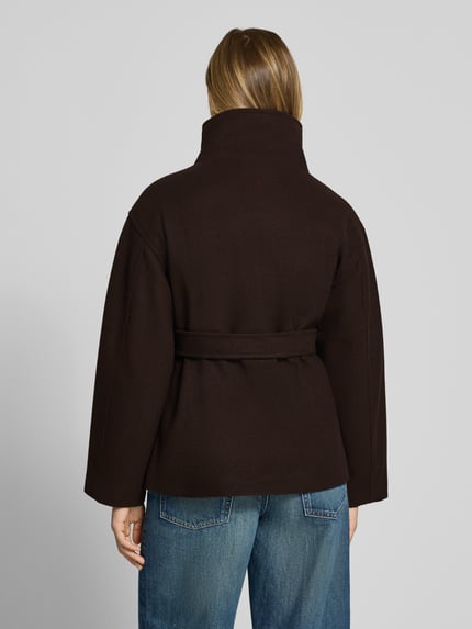 Frauenmodel trägt Jacke mit Stehkragen und Bindegürtel von Gina Tricot in Dunkelbraun, glatte Oberfläche, Ganzkörperansicht Rückansicht. Rückenlänge bei Größe S 70 cm, Schulterbreite 48 cm, Brustweite 48 cm, Ärmellänge 58 cm, Regular Fit, Knopfverschluss, Bindegürtel.