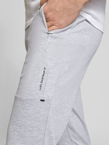 Männermodel trägt sweatpants mit katoenmix Modell 'GORDON' van Jack & Jones in lichtgrijs, ritszakken, elastische band, detailaufnahme rechterzij. Context: heren sweatpants met smalle pasvorm, elastische materiaalmix, tunnelkoord, elastische boorden, zij-ritszakken, binnenbeenlengte maat M 73 cm.