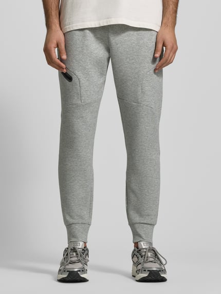 Männermodel trägt Regular Fit Sweatpants mit Logo-Print Modell 'Unstoppable' van Under Armour, elastische materiaalmix met katoen, zak op de pijp met ritssluiting, elastische band en broekspijpuiteinden, binnenbeenlengte maat M: 73 cm, lichtgrijs, ganzkörperansicht von vorne.