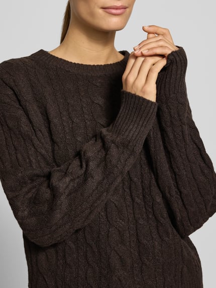 Frauenmodel trägt dunkelbraunen Pullover mit Zopfmuster Modell 'VMSEQUOIA' von VERO MODA, Strick, Rundhals, Nahaufnahme Oberkörper.