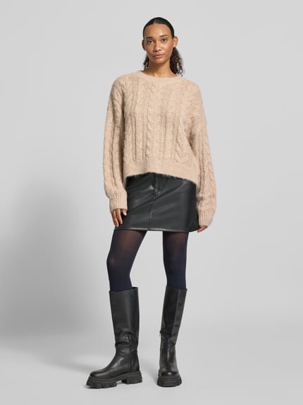 Frauenmodel trägt Oversized Fit Strickpullover mit Zopfmuster von REVIEW in Beige, kombiniert mit kurzem schwarzen Rock, glatter Textur, Ganzkörperansicht von vorne. Rückenlänge S: 54 cm, Ärmellänge S: 58 cm, Rundhalsausschnitt, elastische Abschlüsse.