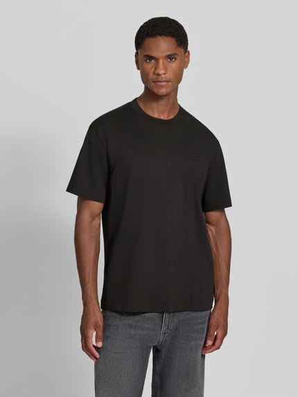 Männermodel trägt Regular Fit T-Shirt aus reiner Baumwolle von Lacoste in Schwarz, kombiniert mit grauer Jeans, Ganzkörperansicht von vorne. Herren T-Shirt von Lacoste