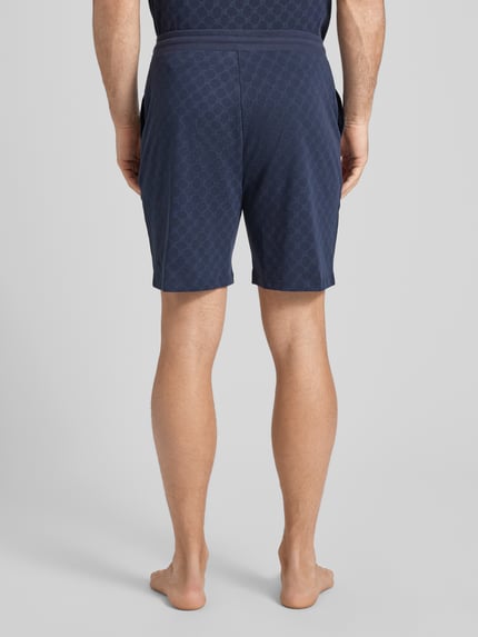 Męski model nosi Shorts aus Baumwolle mit Logo-Muster od JOOP! Collection, czysta bawełna, krój regular fit, elastyczny pas, szerokość w rozmiarze M 42 cm, długość nogawki od wewnątrz w rozmiarze M 18 cm, ciemnoniebieskie, wzór z logo, widok całej sylwetki z tyłu